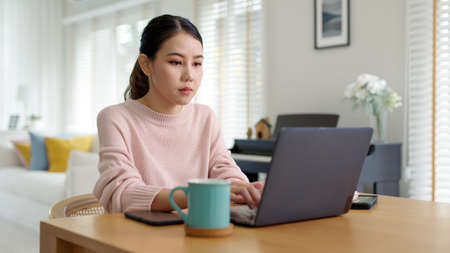 Candid young attractive beautiful asian businesswoman work remotely from home or learning online sitting smiling in cozy workspace at home feeling happy in quarantine social distance concept.の写真素材