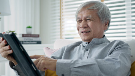 Candid old asian retired guy use tablet computer to transfer money online or financial payment and read text, smile or laugh with happy and positive. Senior asia with digital technology concept.の写真素材