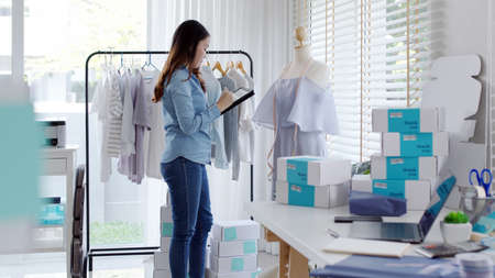 Young attractive beautiful hipster asia female fashion store owner use digital tablet check stock clothes or package box on rail rack at home office small SME business entrepreneur e-commerce concept.の写真素材