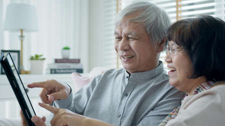 Candid old asian retired couple use tablet computer to transfer money online or financial payment and read text, smile or laugh with happy and positive. Senior asia with digital technology concept.の写真素材