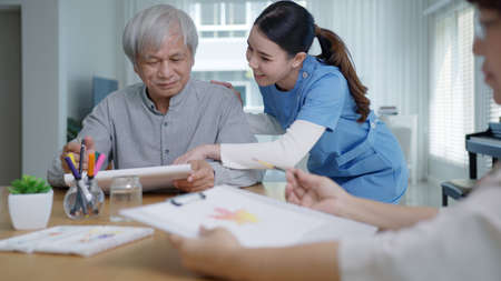 Attractive young senior asian citizen couple sit at home watercolor painting artwork on desk table with nurse feeling happy in mental health therapy or asia older people quarantine activity lifestyle.の写真素材