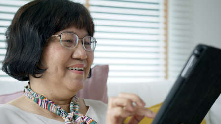 Candid old asian retired lady use tablet computer to transfer money online or financial payment and read text, smile or laugh with happy and positive. Senior asia with digital technology concept.の写真素材