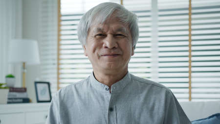 Portrait of happy senior asian man relax sitting smile at home apartment on sofa couch looking at camera with enjoy retirement or quarantine time. Attractive asia adult older people happy lifestyle.の写真素材