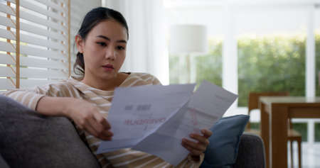 Upset tired young asia sad lady people sit sofa couch at home think hard worry in tax expense saving...