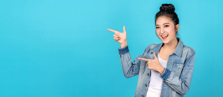 Banner of Happy asian woman standing pointing hands to copyspace on blue background. Cute asia girl smiling wearing casual jeans shirt and finger pointing to aside for present promotions.の写真素材