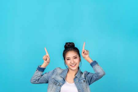 Happy asian woman feeling happiness and standing pointing hands up to copyspace on blue background.Cute asia girl smiling wearing casual jeans shirt and finger pointing to above for present promotionsの写真素材