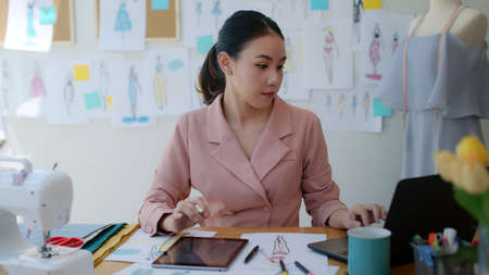 Young happy smile asia success woman busy work on new dress collection on desk laptop tablet at...