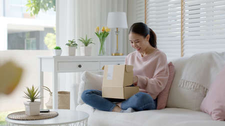 Young happy asia people teen girl smile enjoy unbox post mail sit relax at home comfort sofa couch feel joy amazed awe wow in omni channel fast send parcel via online sale shop store e-commerce order.の写真素材