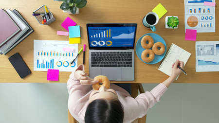 Above top view of asia girl or woman staff worker sitting busy work in multi task job or relax study MBA at home office on desk eat bite diet cake fast food late lunch meal in hurry break coffee time.の写真素材