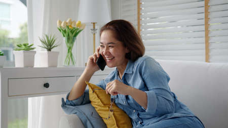Asia elder lady people, latin mom sitting at home cozy sofa couch use digital 5G cellular cellphone on wifi wireless service support in remotely social distance call to advice, gossip, order online.の写真素材