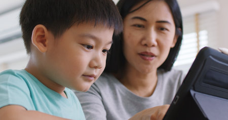 Cute asia people alpha Gen Z kid small boy play video game app online happy mum at home. Enjoy Child care fun class little son and mom study learn upskill idea course on smart digital tablet computer.の写真素材