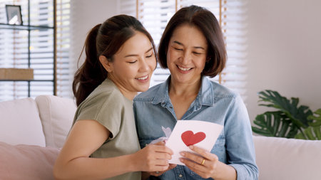 May Mother's day young adult grown up child cuddle hug give flower gift box red heart card to mature middle aged mum. Love kiss care mom asia people sitting at home sofa happy smile enjoy family time.の素材 [FY310213329320]