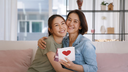 Happy time Mother day grown up child girl looking at camera cuddle hug give gift box heart card to mature mum. Love kiss care mom asia middle age adult people smile enjoy relax sitting at home sofa.の写真素材