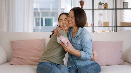 May Mother's day young adult grown up child cuddle hug give flower gift box red heart card to mature middle aged mum. Love kiss care mom asia people sitting at home sofa happy smile enjoy family time.の写真素材