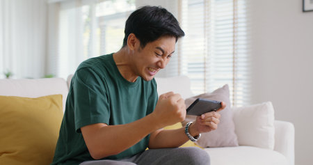 Asia people young man relax sitting easy at home sofa couch laugh smile enjoy play video game social media on mobile app, win online stream casino battle trade, leisure teen activity happy fun joy.の写真素材