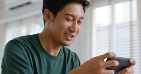 Asia people young man relax sitting easy at home sofa couch laugh smile enjoy play video game social media on mobile app, win online stream casino battle trade, leisure teen activity happy fun joy.の写真素材