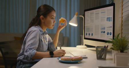 Asia people young girl work busy at home office sit on desk crave sweet junk fast food eat bite donut at night. Workforce issue bad habit stress relief by fat diet break time late dinner in hard job.の写真素材