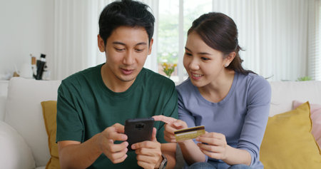 Secure pay gift card spend money buy ticket order from retail store on phone social media app. Asia people happy man woman, husband wife young couple sit at home sofa enjoy fun talk smile laugh relax.の写真素材