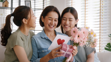 Mother day two grown up child cuddle hug give flower gift box red heart card to mature mum. Love kiss care mom asia middle age adult three people sitting at home sofa happy smile enjoy family time.の写真素材