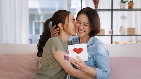 May Mother's day young adult grown up child cuddle hug give flower gift box red heart card to mature middle aged mum. Love kiss care mom asia people sitting at home sofa happy smile enjoy family time.の写真素材