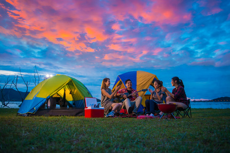 Camping of happy asian young travellers at lake, asian man and women group, relaxing, sing a song and cooking, at sunset.の写真素材