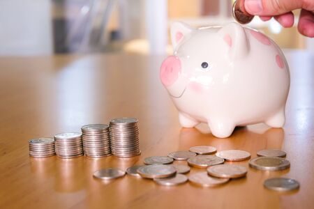 Selective focus of Men hand putting coin to piggy bank with stack of coin on wooden desk fill with natural light background. Saving money and financial, life expense planning lifestyle wealth concept.の写真素材