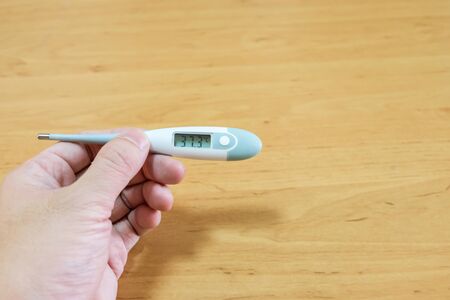 Men hand holding digital thermometer to measure body temperature and check for fever on wood background. Sickness, healthcare and medical concept.の写真素材