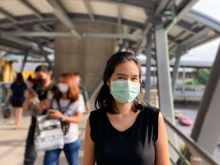 Adult Asian Thai woman wearing surgical face mask on public sky train walkway. With many people blurred in the background. People need to prevent and protect from infect from Coronavirus. New normalの写真素材