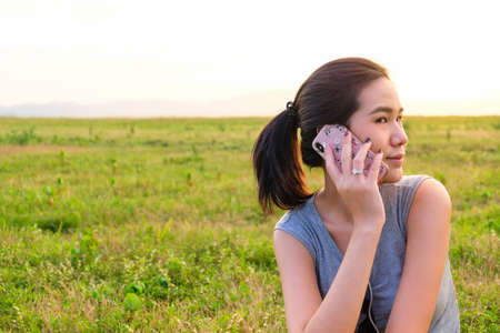 Selective focus of Asian women taking on mobile phone with green grass and sunset nature backgroundの写真素材
