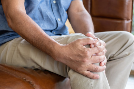 Selective focus at hand of old senior Asian man sitting at sofa touching knee and suffer from the pain and injury. Healthcare and medical problem of Elder people concept.の写真素材