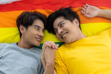 Asian gay homosexual couple holding hands on the bed.  With rainbow flag as LGBT sign at the background. Gender equality and right concept, playful and romantic moment.の写真素材
