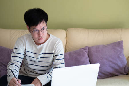 Adult Asian men sitting at the sofa couch while using laptop and write down the note on the paper. Online learning or work from home concept to promote social distancing, new normal after Covid-19の写真素材