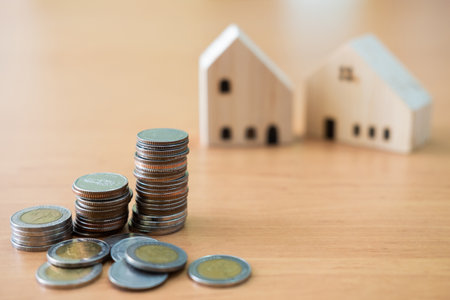 Selective focus at increase coins stack. With blurred house mock up on wooden table as background. Saving plan for home ownership or real estate buying, loan concept.の写真素材