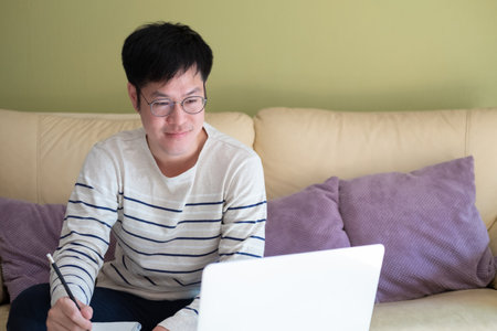 Adult Asian men sitting at the sofa couch while using laptop and write down the note on the paper....
