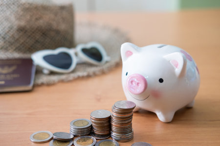 Selective focus at piggy bank. Stack of money coin with blurred straw hat, sunglasses and passport mock up at the background. Money saving plan for holiday or vacation concept.の写真素材
