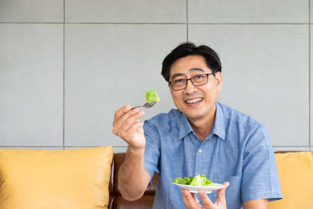 Senior Asian men sitting on the sofa at home while eating green vegetable salad and organic orange juice for breakfast. Healthy eating lifestyle for Elder people concept.の写真素材