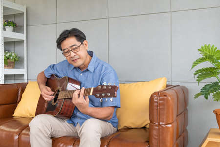 Asian elder man enjoy playing guitar at the sofa inside of well interior decoration house. Active senior lifestyle after retirement with happiness with a good mental health.の写真素材