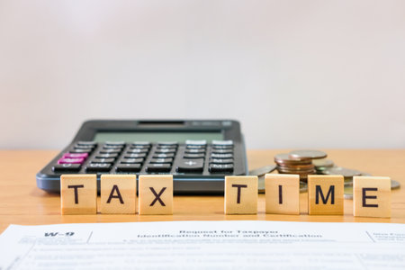 Selective focus at wood tax time sign with blurred calculator and coins stack and white copy space background. Business or personal tax payment, expense that need to manage and plan.の写真素材