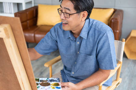 Indoor shot of Senior Asian man sit and painting picture on easel inside of the house. Leisure activity for elderly for relaxation,  happy retirement lifestyle concept.の写真素材