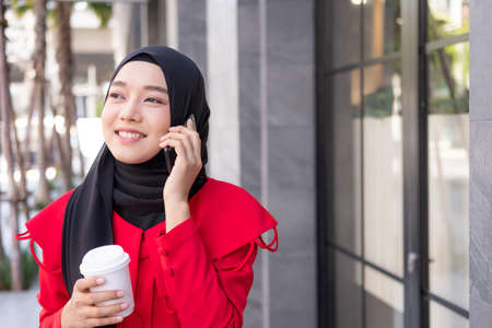 Beautiful East Asian Islamic women wearing hijab with formal dress. Talking on mobile phone while holding coffee cup and walking in urban city area or department store. People lifestyle concept.の写真素材