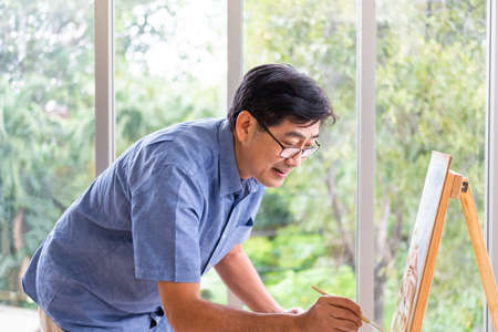 Indoor shot of senior Asian men using brush and oil color to paint on painting canvas. With smile on...