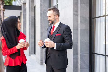 Selective focus at men face. Outdoor shot of Smart Business man and women in formal suit holding coffee cup while standing at Central Business district in the. Diverse people working environment.の写真素材