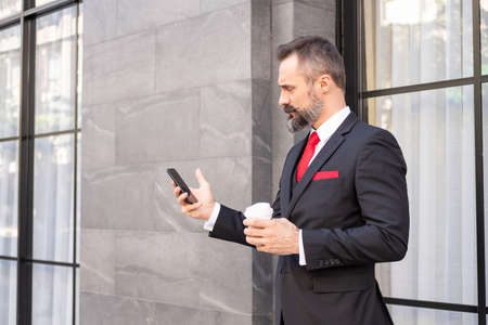Selective focus at face. Adult Caucasian business man with beard, dress in formal suit standing in central business district. While drink morning coffee and look serious at his mobile smartphone.の写真素材