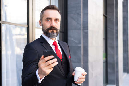Selective focus at face. Adult Caucasian business man with beard, dress in formal suit standing in central business district. While drink morning coffee and look serious at his mobile smartphone.の写真素材