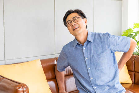 Indoor shot of senior Asian men sit on the sofa couch and touch his back and suffering from back pain, aching muscles, bones or lumbago. Elderly health problem concept.の写真素材