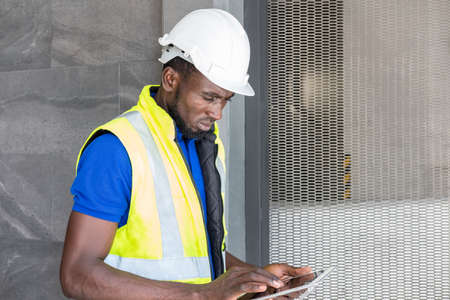 Selective focus at face of Black African foreman at building construction site, wearing protective hat and safety equipment while using digital tablet to record information. Civil engineer working.の写真素材