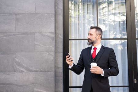 Selective focus at face. Adult Caucasian business man with beard, dress in formal suit standing in central business district. While drink morning coffee and look serious at his mobile smartphone.の写真素材