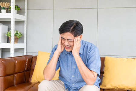 Normal shot of Asian male senior using hands to touch his head when suffering and pain from headache. Health problem or stress in elder people. Healthcare and medical concept.の写真素材