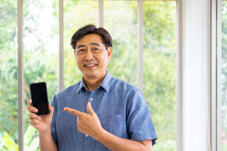 Asian elder man holding smartphone mock up with empty screen to add logo of application while point at that device with smile. Active senior  and technology with blurred green nature background.の写真素材