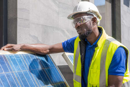 Outdoor shot of black African engineer inspect electrical solar panel wearing hardhat , protective eyeglass and safety equipment with smile on his face. Alternative energy and industrial job concept.の写真素材